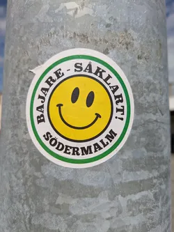 Street sticker The sticker is circular with a yellow smiley face in the center. The text "BAJARE - SÅKLART!" is on the top of the sticker and "SÖDERMALM" is at the bottom, both written in black and set against a green background.