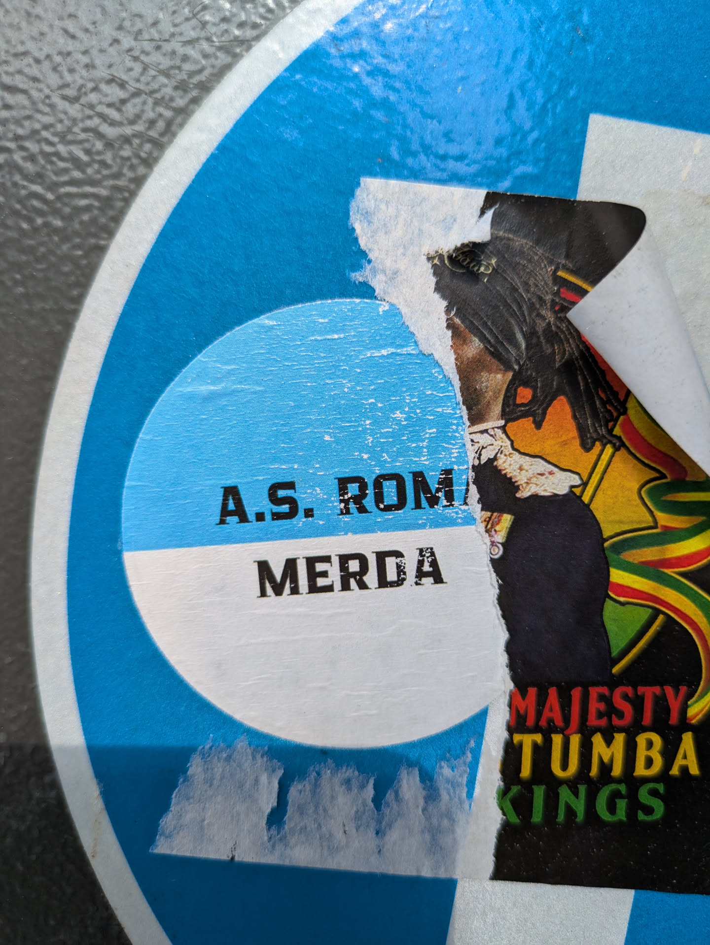 The sticker is heavily torn and weathered, revealing two distinct visual sections. The left section is part of a circular design in blue and white, featuring bold black text that reads "A.S. ROMA" and "MERDA" (Italian for "shit"). The right section, which is significantly ripped, depicts a stylized figure with dark dreadlocks, wearing what appears to be a dark uniform with medals. Behind this figure, there's a vibrant background with flowing ribbons or banners in red, yellow, and green, characteristic of Rastafarian colors. Below the figure, partially visible text in red, yellow, and green lettering spells out "MAJESTY", "TUMBA", and "KINGS". The overall appearance suggests a blend of football fan culture and music-related art.