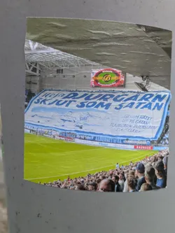 Street sticker A sticker depicting a soccer stadium scene with a large banner reading "Heja Djurgården Skjut som Satan" (Go Djurgården! Shoot like Satan). The banner also includes additional text in Swedish. The sticker shows a lively match atmosphere with spectators in the stands.