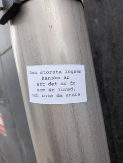 A rectangular white paper sticker with black text printed in a typewriter-style font. The sticker is adhered to a brushed metallic cylindrical surface, possibly a pole or a container. The edges of the paper appear slightly worn or curled, indicating it's a physical object. The text is centered on the sticker.