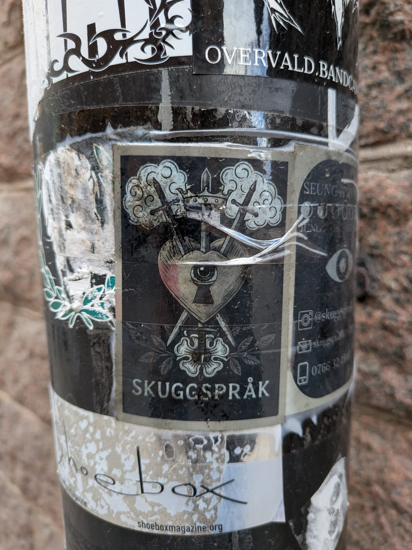 The central sticker is a rectangular design with a dark, possibly black or very dark grey, background. Its main motif is a stylized heart, subtly tinted with pink or red, which contains a keyhole and an eye looking through it. Two ornate swords cross behind the heart, their hilts visible above. A small crown rests atop the heart and swords. Above the crown, there are swirling cloud-like elements. Below the heart, two symmetrical floral designs, resembling roses or similar blossoms, are depicted with leaves. The overall style is intricate and gothic. At the very bottom of this central panel, the word 'SKUGGSPRÅK' is written in a clear, sans-serif font. To the right, on an adjacent panel that appears to be part of the same sticker or a closely related one, there are icons for Instagram, email/website, and phone, along with corresponding text.