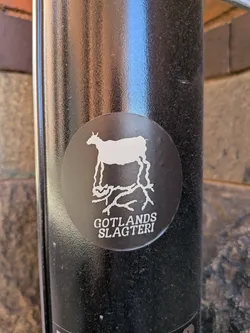 Street sticker The sticker is circular with a dark background and a white silhouette of a goat. The goat is depicted in profile, standing on what appears to be stylized roots or branches. Below the goat's legs, in white capital letters, is the text 'GOTLANDS SLAGTERI'. The sticker appears to be adhered to a textured, dark cylindrical object.