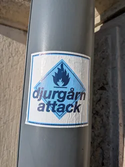 Street sticker The sticker is rectangular with a white background and a blue border. At the center, there is a blue diamond shape containing a stylized blue flame symbol. Below the flame, the text 'djurgårn attack' is written in large, dark blue, sans-serif capital letters. At the very bottom of the diamond, in smaller text, are the words 'biaränderna'. The sticker appears slightly creased and aged, suggesting it is a real sticker applied to a surface.