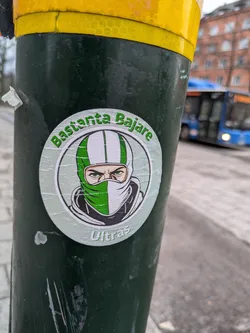 Street sticker A round sticker featuring a person wearing a green and white balaclava. The text 'Bastanta Bajare' is written in a circle around the image, and the word 'Ultras' is written below it.
