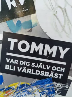 Street sticker A black and white sticker with the word TOMMY at the top and the Swedish phrase "Var dig själv och bli världsbäst" (Be yourself and become the best in the world) below it.