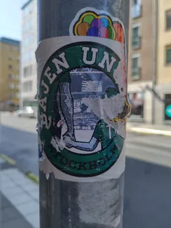 A round sticker featuring an illustration of a person, possibly an athlete, with text that reads "BASEN UNITE STOCKHOLM." The sticker is affixed to a metal pole and shows some wear and tear. Part of the central image is obscured. 