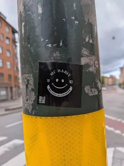 A square black sticker with white text and a white smiley face, affixed to a dark green, weathered pole. The top text reads 'MC HABIT', with '19' and '83' flanking the smiley face. Below the smiley, an arc of text reads 'MÄSTAREN AV CEREMONIN'. A small, white QR code is visible in the bottom left corner of the sticker.