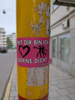 Street sticker The sticker is rectangular with a vibrant pink background. On the left, there is a large, roughly drawn black heart. To its right, two black stick figures are depicted, one holding a beer mug and the other a spray can or bottle. White, sans-serif capital letters read "MIT DIR BIN ICH" above the heart and figures, and "GERNE DICHT" below them. At the very bottom right, a small Instagram logo is followed by "@AGRARSTICKER". The sticker shows some minor wear, particularly around its edges, and is affixed to a reflective yellow pole.