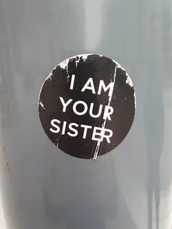 Street sticker A round black and white sticker with the text "I AM YOUR SISTER" on it. The sticker is attached to a gray surface and is slightly damaged.
