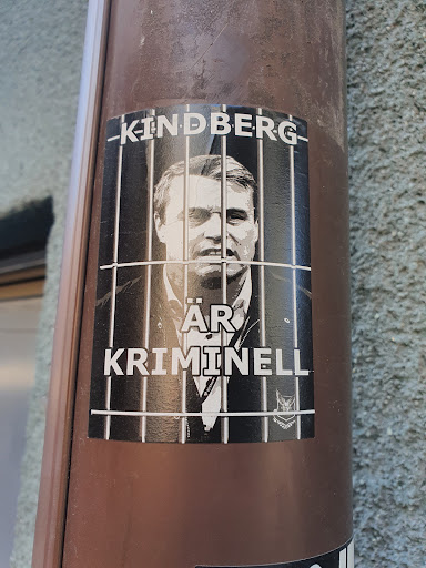 Street sticker Black and white sticker depicting a man behind bars with the text "Kindberg är kriminell" (Kindberg is a criminal) in Swedish. The sticker has a logo at the bottom right corner resembling a stylized bird of prey.