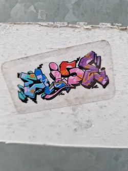 A rectangular sticker featuring stylized graffiti lettering that spells out the word "WISS". The letters are in various shades of pink, purple, blue, and orange with added details and splatters. The sticker is affixed to a light gray surface.