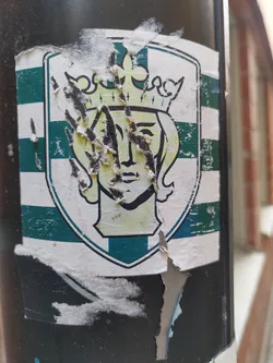 This sticker shows a worn-out emblem with a woman wearing a crown inside a shield-shaped design.  The colors are mainly green and white. The sticker is significantly damaged and peeling.