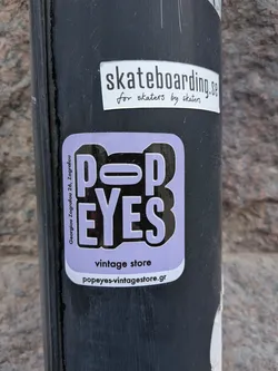 Street sticker A light lavender, rounded rectangular sticker featuring the bold, blocky text 'POP EYES' in a slightly darker shade of purple with a prominent black outline, creating a 3D effect. The 'O' in 'POP' and the 'E' in 'EYES' incorporate horizontal line elements within their letterforms. Vertically aligned on the left side, in smaller white text, is 'Georgiou Zografou 26, Zografou'. Below the main logo, in black text, it reads 'vintage store', and at the very bottom, 'popeyes-vintagestore.gr' is displayed.