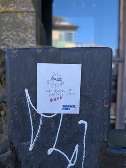 Street sticker A white, rectangular sticker featuring a child-like stick figure drawing of a superhero with a blue eye mask and a wide smile, arms raised in a triumphant pose. Below the drawing, the Swedish text 'alla barn är superhjältar' (all children are superheroes) is written in black, followed by three small red hearts. In the bottom right corner, the logo 'BARNCANCER FONDEN' (Child Cancer Fund) is printed in blue text. The sticker has a slightly worn appearance and is affixed to a dark, textured metal post with white graffiti underneath.