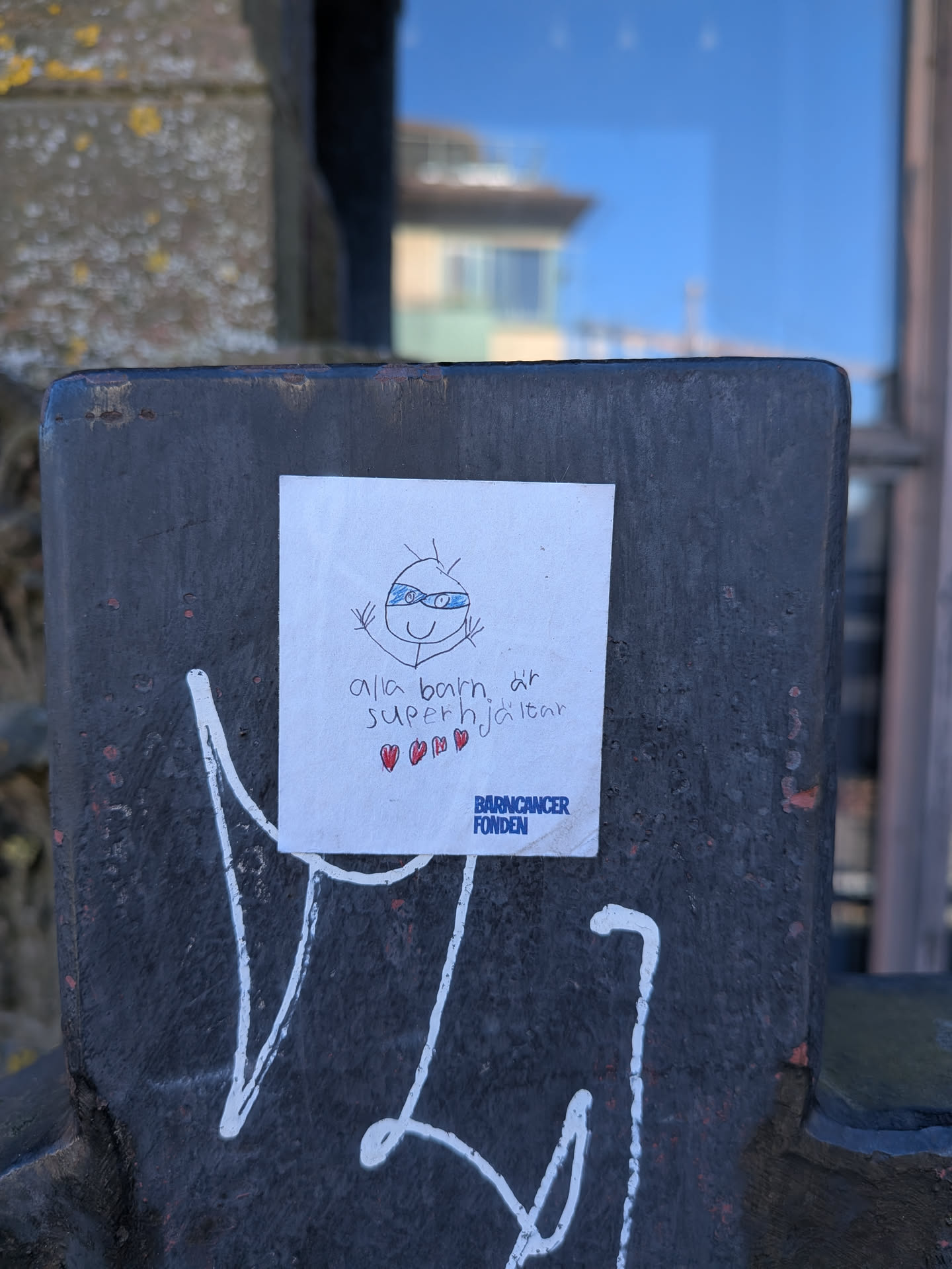 A white, rectangular sticker featuring a child-like stick figure drawing of a superhero with a blue eye mask and a wide smile, arms raised in a triumphant pose. Below the drawing, the Swedish text 'alla barn är superhjältar' (all children are superheroes) is written in black, followed by three small red hearts. In the bottom right corner, the logo 'BARNCANCER FONDEN' (Child Cancer Fund) is printed in blue text. The sticker has a slightly worn appearance and is affixed to a dark, textured metal post with white graffiti underneath.