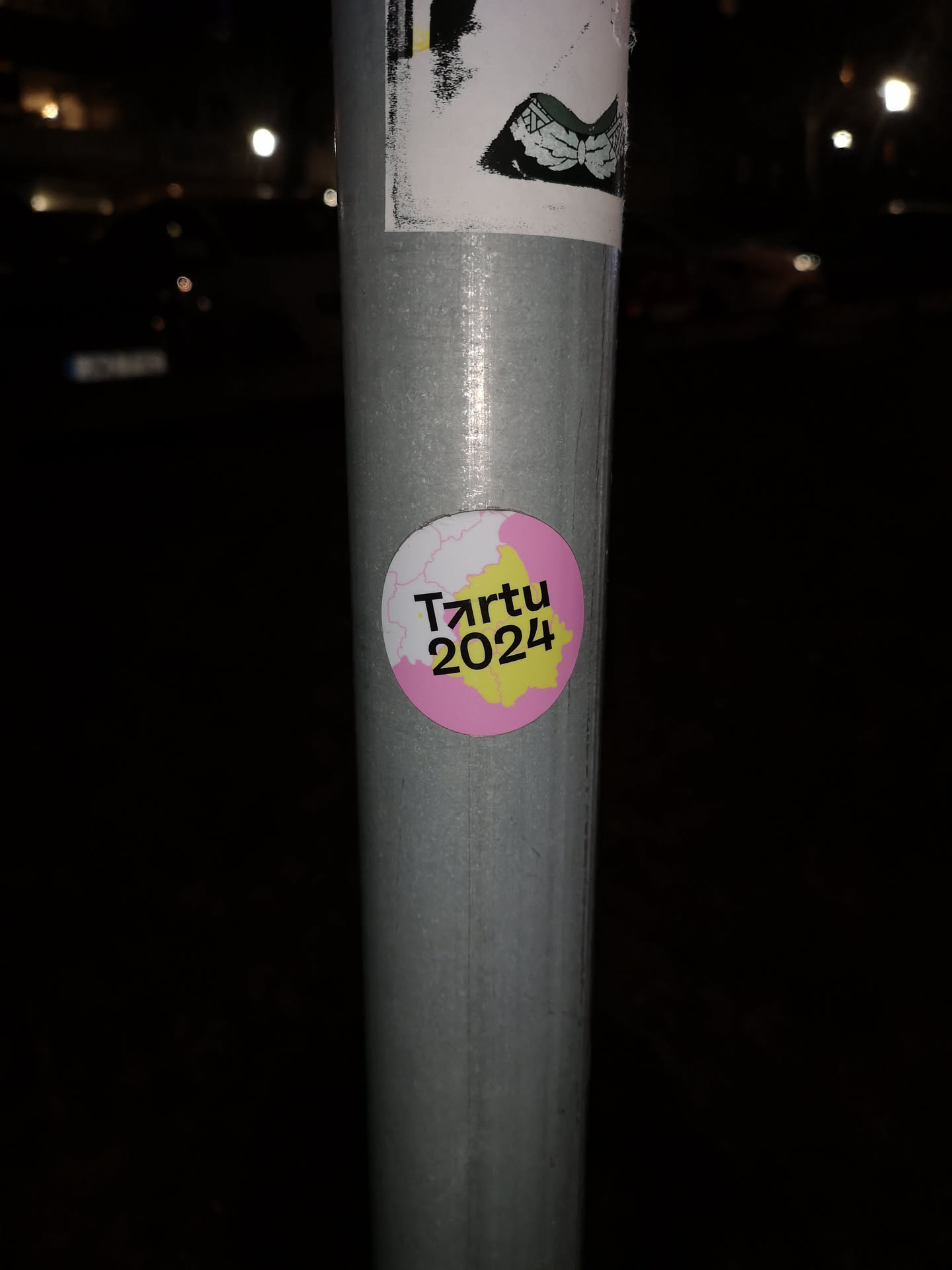 A circular sticker featuring a vibrant design with a pink background on the left and a bright yellow background on the right, separated by a white, irregular, map-like outline. The text 'Tartu' is prominently displayed in black, with the 'r' stylized as an upward-pointing arrow. Below 'Tartu' is the year '2024' in black. The sticker has a clean, modern aesthetic, promoting the city of Tartu and its cultural year.
