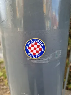 Street sticker A round sticker featuring the logo of Hajduk Split, a Croatian football club. The logo includes the club's name and a checkered pattern reminiscent of the Croatian flag.  The sticker is affixed to a gray pole.