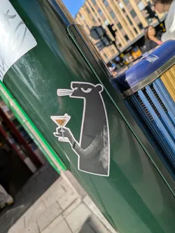 Die-cut sticker depicting a stylized black animal in profile. The character has a nonchalant expression, holds a martini glass with an olive, and has smoke lines coming from its mouth. It is outlined in white.