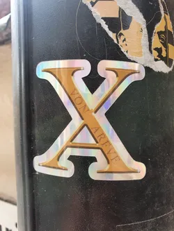 A holographic sticker with a gold letter X. The word "Von Arve" is written vertically inside the letter. The sticker has an iridescent finish that shifts color in different lights.