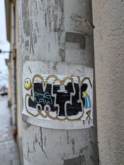 Street sticker The sticker is rectangular, predominantly white, and affixed to a weathered grey pole. Its central feature is a large, stylized graffiti tag rendered in thick black fill with a prominent white outline, further bordered by a wavy, metallic gold/bronze outline. Within the black fill of the graffiti, smaller, light blue/teal text is visible. To the left of the main graffiti, there's a bright yellow circular smiley face with black eyes and a smile. To the right, a small, cartoonish shooting star is depicted with a yellow star and a blue and white striped tail. The sticker shows some signs of wear and slight creasing, indicating it's a physical object on an outdoor surface.