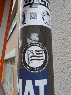 Street sticker The sticker is circular, predominantly black and white. It features a stylized flag with horizontal stripes and a prominent 'ST' monogram in the upper left canton, waving on a flagpole. The flag is centered within a white circle. Around the inner edge of the black border, the text 'SK STURM GRAZ' is written at the top, and 'SEIT 1909' is written at the bottom, separated by small dots on either side. The overall design is a classic club emblem.