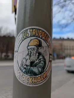This is a circular sticker on a gray pole. The sticker's design features a person with a long beard wearing a yellow hardhat and a dark coat. The text "JUL TRUPPEN" is at the top and "STOCKHOLM" is at the bottom. The number "23" is also visible on the right side of the design.