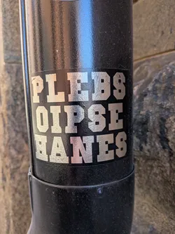 Street sticker The sticker features the text "PLEBS OIPSE HANES" in large, bold, block-style letters. The letters are silver with a slightly distressed or textured finish, appearing almost metallic. They are arranged in three rows, with the first row containing "PLEBS", the second row "OIPSE", and the third row "HANES". The sticker has a black background, and its edges appear to be slightly worn or rounded, suggesting it is a peel-and-stick label applied to a curved black surface, possibly a pipe or a cylindrical object.