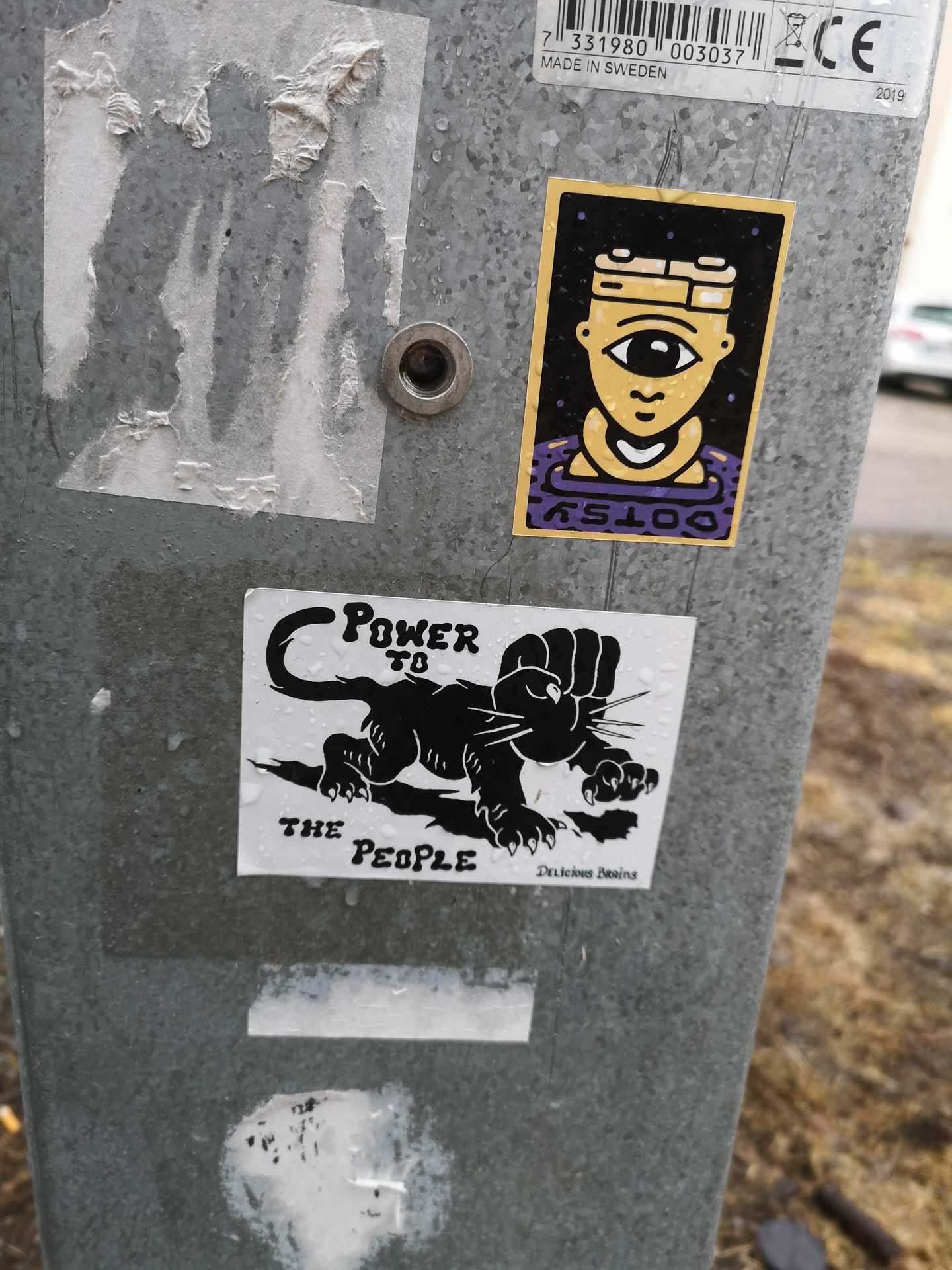A monochrome sticker featuring a stylized black panther in a crouching, ready-to-pounce stance. The panther's right front paw is replaced by a clenched human fist, raised in a symbol of power and solidarity. The text 'POWER TO' is positioned above the panther's back, and 'THE PEOPLE' is below its front paws. The design is black on a white background, with some visible water droplets on its surface. In the bottom right corner, smaller text reads 'Delicious Brains'.