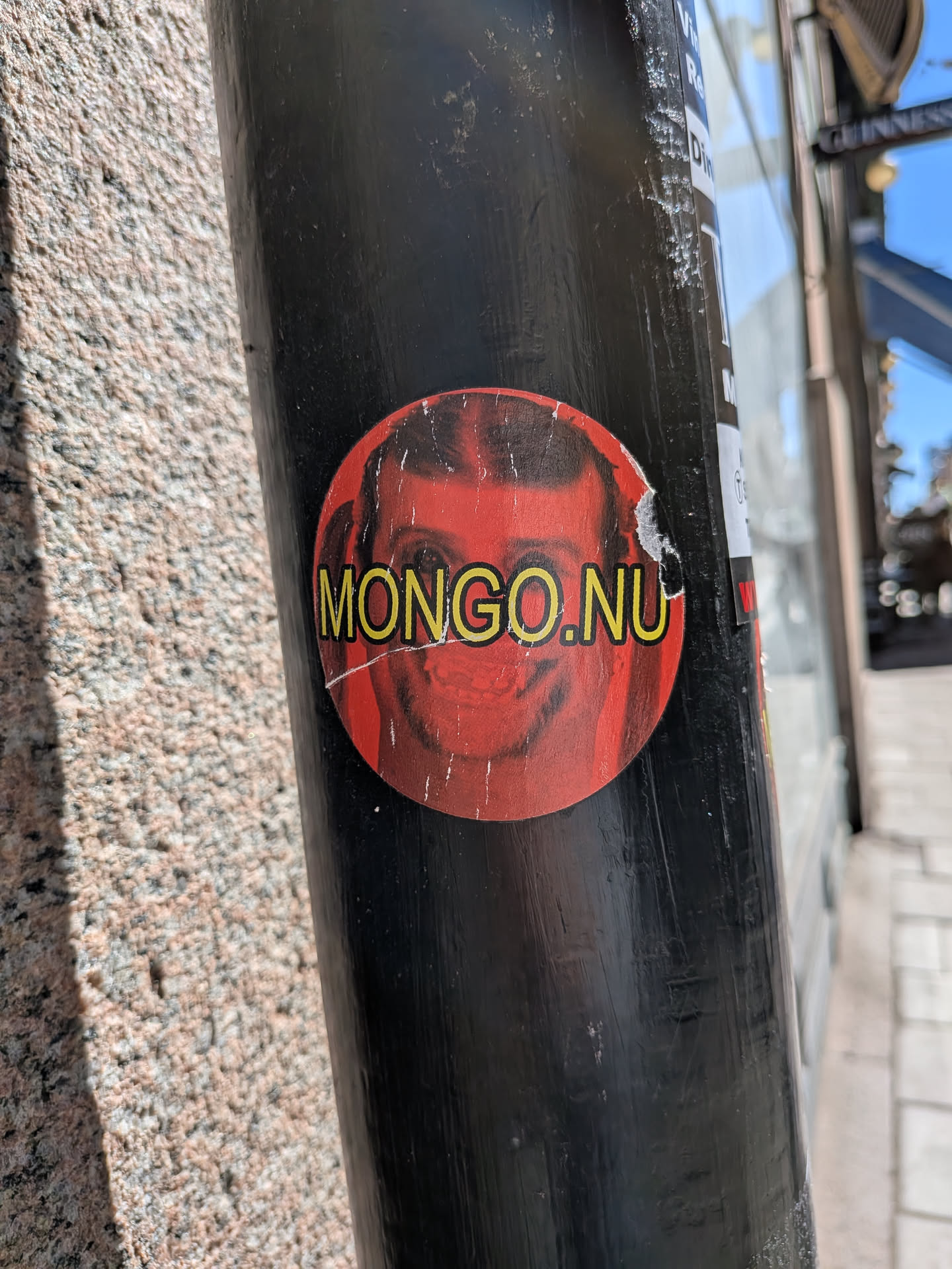 The sticker is circular, featuring a striking red background. Superimposed on this red is a stylized, somewhat distorted image of a man's face, rendered in darker, almost shadowy tones, with prominent eyes and a subtle, intense expression. Across the center of the face, in bold yellow capital letters, is the text "MONGO.NU". The sticker shows visible signs of wear, including white scuffs and minor peeling around its edges, indicating its exposure to the elements. It is affixed to a black pole.