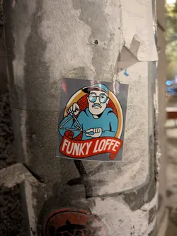 Street sticker The sticker features a cartoon illustration of a man wearing glasses, a blue hat, and a blue shirt. He has a mustache and is holding a musical triangle with a beater. The illustration is enclosed in an oval with a red and white dotted background and an orange border. Below the oval, a red banner with white text reads 'FUNKY LOFFE'. The overall style is reminiscent of retro cartoons.