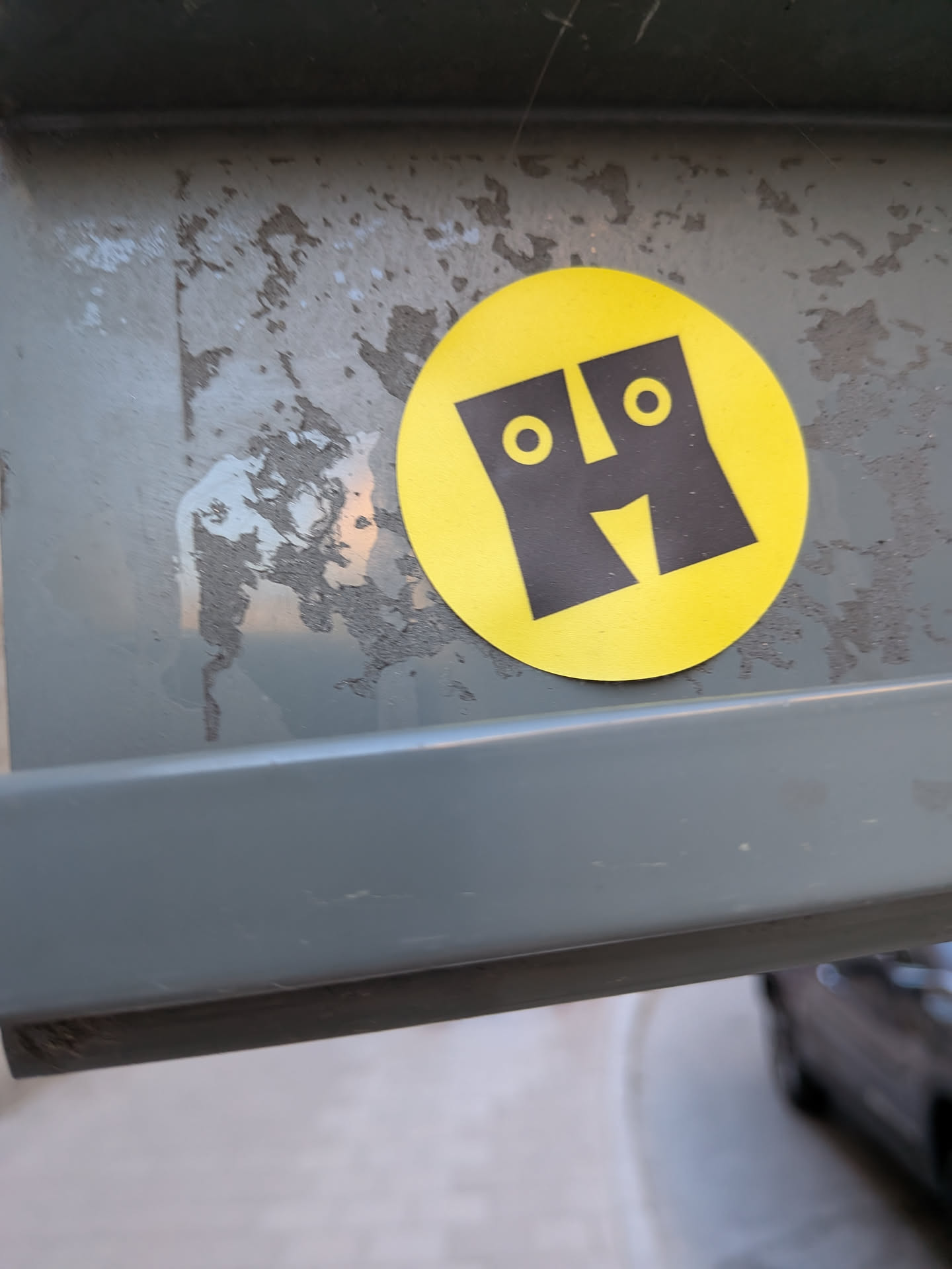 The sticker is circular with a vibrant yellow background. At its center is a bold, black, stylized representation of the letter 'H'. The vertical strokes of the 'H' are wide and rectangular. Within the upper portion of each vertical stroke, there is a small, white-ringed black circle, serving as eyes. The horizontal bar of the 'H' is replaced by a downward-pointing 'V' shape, which creates the impression of a nose or a smiling mouth. The overall design is highly graphic and minimalist, transforming the letter 'H' into a cheerful, anthropomorphic character.