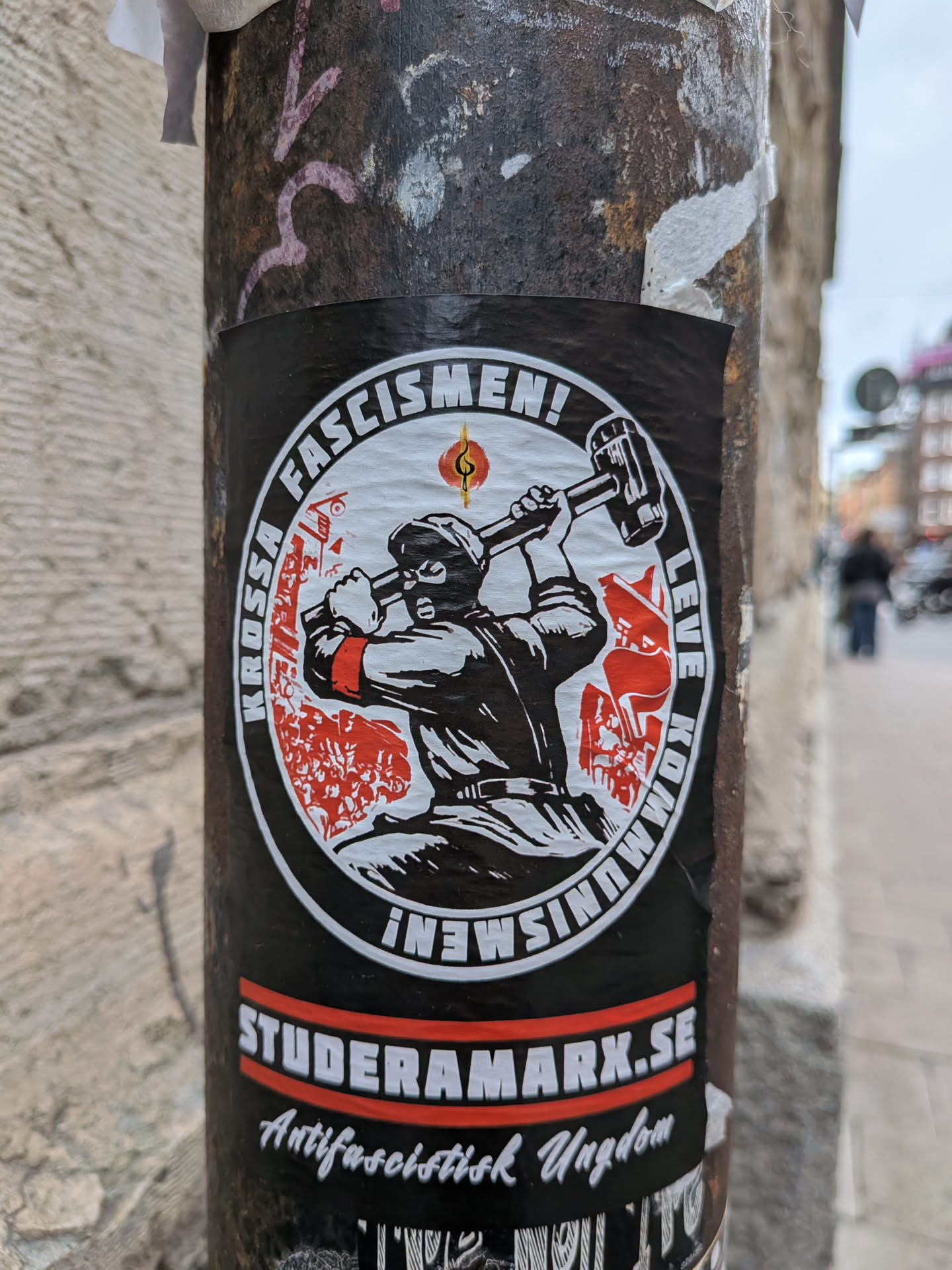 A black rectangular sticker featuring a central circular emblem. Inside the circle, a black-and-white illustration depicts a masked individual wearing a red armband and wielding a large sledgehammer. The background of the emblem shows red silhouettes of a crowd and a flag. The circular border contains the Swedish text 'KROSSA FASCISMEN! LEVE KOMMUNISMEN!'. Below the circle, a red bar displays the website 'STUDERAMARX.SE', followed by the script text 'Antifascistisk Ungdom'.