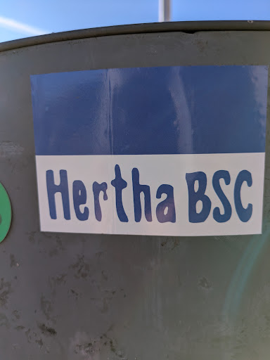 The sticker features the Hertha BSC logo with a blue and white color scheme. The text 'Hertha BSC' is prominently displayed in a rounded, blue font on a white background. The top portion of the sticker is a solid blue. The sticker is affixed to a slightly weathered, grey metal surface.