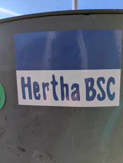 Street sticker The sticker features the Hertha BSC logo with a blue and white color scheme. The text 'Hertha BSC' is prominently displayed in a rounded, blue font on a white background. The top portion of the sticker is a solid blue. The sticker is affixed to a slightly weathered, grey metal surface.