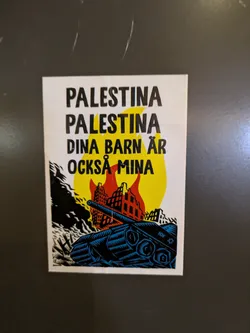 Street sticker The sticker depicts a blue tank in the foreground, with burning buildings against a yellow backdrop. The text "Palestina Palestina Dina Barn Ar Ocksa Mina" is displayed at the top. The sticker uses a limited color palette and blocky style.