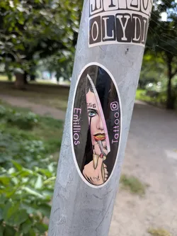 Street sticker Oval sticker depicting a woman with a '666' forehead tattoo holding a knife reflecting her face. Flanked by pink text 'Emillios' and 'Tattoo' with an Instagram icon.