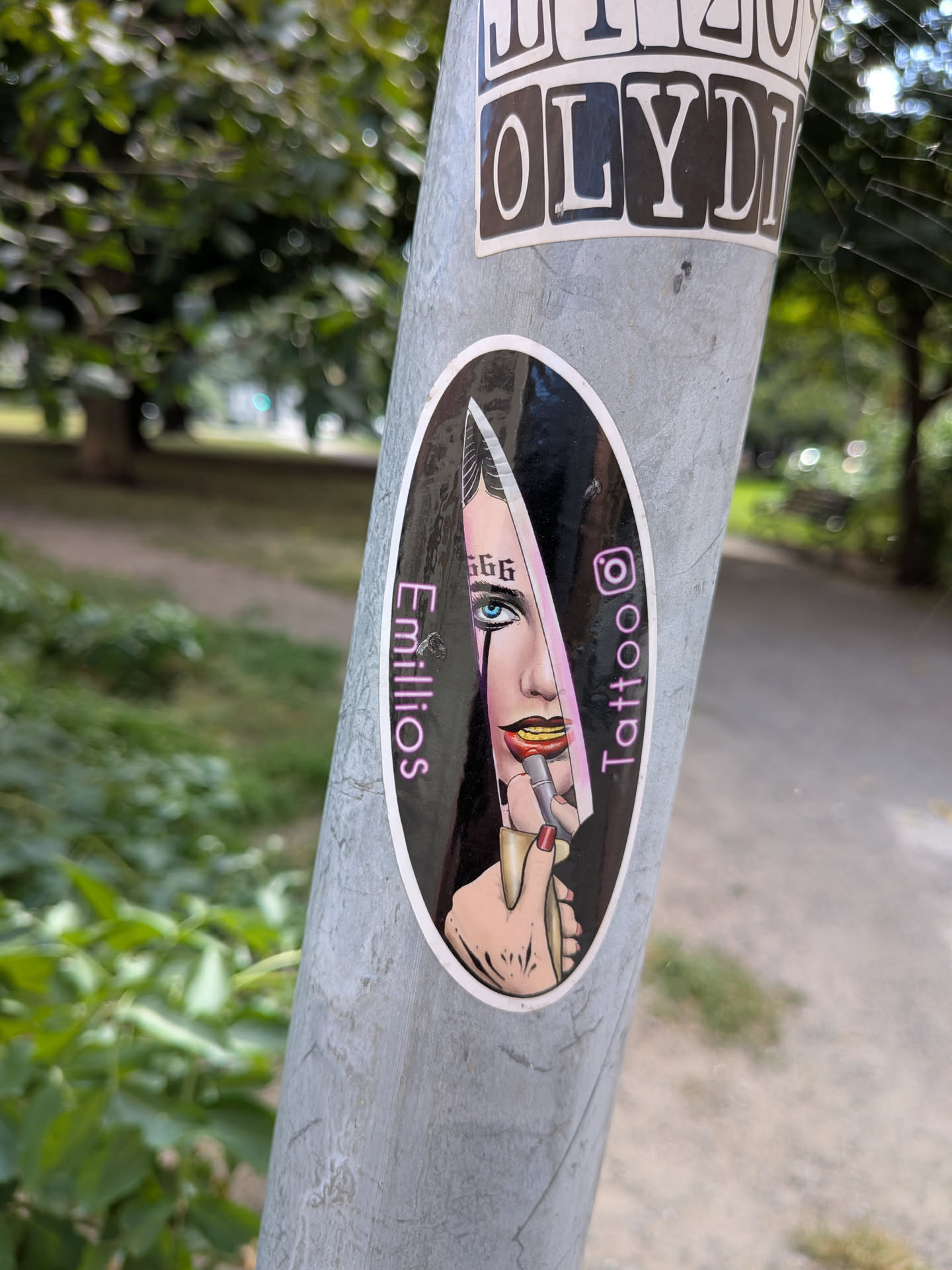 Oval sticker depicting a woman with a '666' forehead tattoo holding a knife reflecting her face. Flanked by pink text 'Emillios' and 'Tattoo' with an Instagram icon.