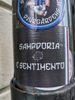 Street sticker Black and white sticker with the text "Sampdoria è sentimento" and a stylized wolf's head in between.  The sticker is on a pole with other stickers around it.