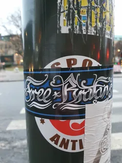 Street sticker A sticker with the text "Free Furryans" in silver stylized lettering on a dark background with a blue stripe at the bottom.  The sticker is affixed to a pole, among other stickers.