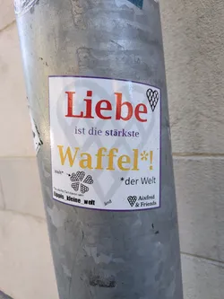 Street sticker A rectangular white sticker with a thin purple border, adhered to a galvanized metal pole. The main text, in German, reads 'Liebe ist die stärkste Waffel*!' ('Love is the strongest waffle!'). 'Liebe' is in large red serif font, 'ist die stärkste' in smaller purple font, and 'Waffel*!' in large yellow serif font. A faint grey waffle pattern is visible in the background of the text. Decorative elements include a grid-patterned heart outline in the top right corner and a clover-like arrangement of four grid-patterned hearts on the left side. Smaller text at the bottom includes 'Welt*', '*der Welt', 'Du darfst Teil davon sein', 'Lippis_kleine_welt' (in bold black), 'and', and 'Aixfred & Friends' next to a small grid-patterned heart.