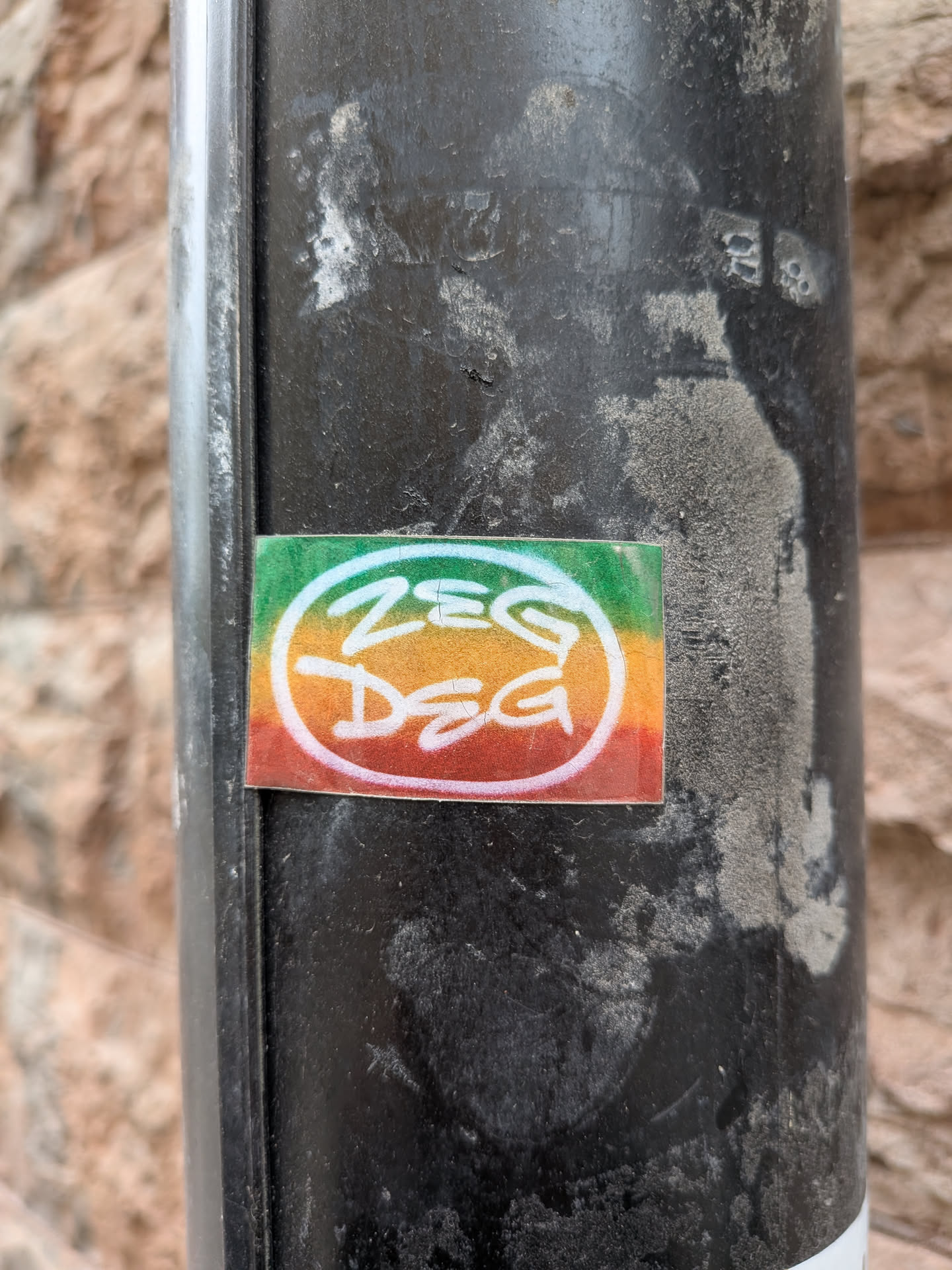 A rectangular sticker featuring a horizontal gradient background in the iconic Rasta colors: green on the left, transitioning to yellow/gold in the center, and red on the right. Overlaid on this colorful backdrop is white, stylized text that reads 'ZEG DEG'. The word 'ZEG' is positioned above 'DEG', both rendered in a thick, rounded, graffiti-like font with a slightly distressed or hand-drawn appearance. The sticker has a glossy finish and is adhered to a dark, weathered metal pole.