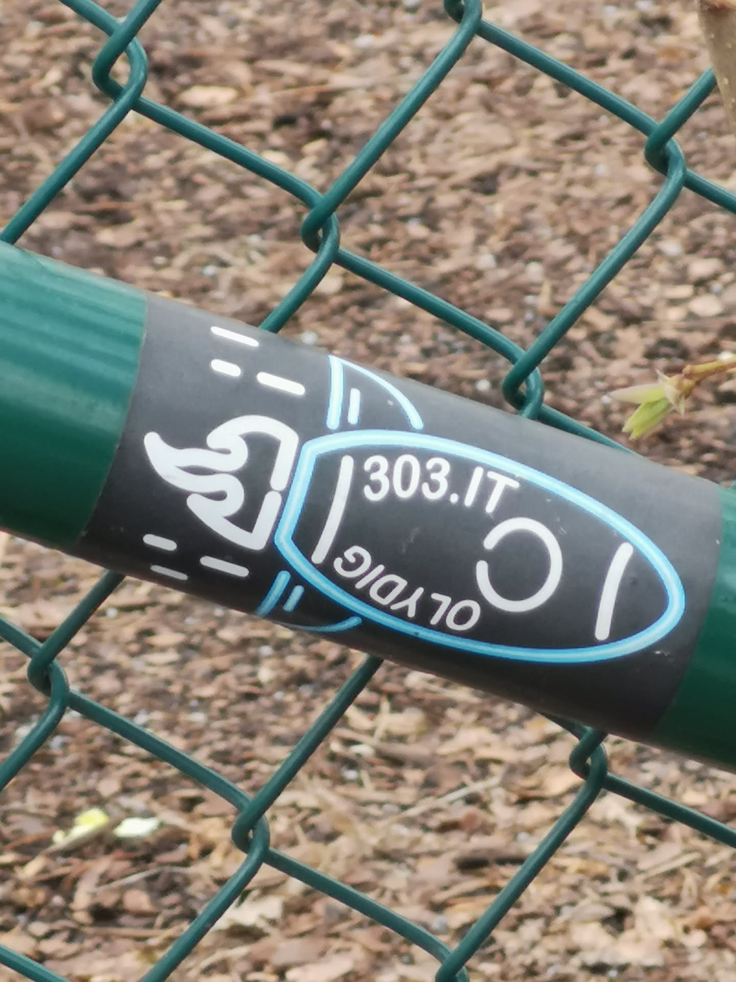The sticker is rectangular, predominantly dark grey or black, wrapped around a green chain-link fence post. On the left, there's a stylized, minimalist outline of a white duck or bird head facing left, with white dashed lines extending horizontally above and below it. To the right, an oval shape is outlined in bright light blue and white. Inside this oval, the text '303.IT' is prominently displayed horizontally in white. Below it, the word 'OLYDIG' is written in white, curved along the bottom edge of the oval and appears upside down relative to '303.IT'. The overall aesthetic is clean and graphic.