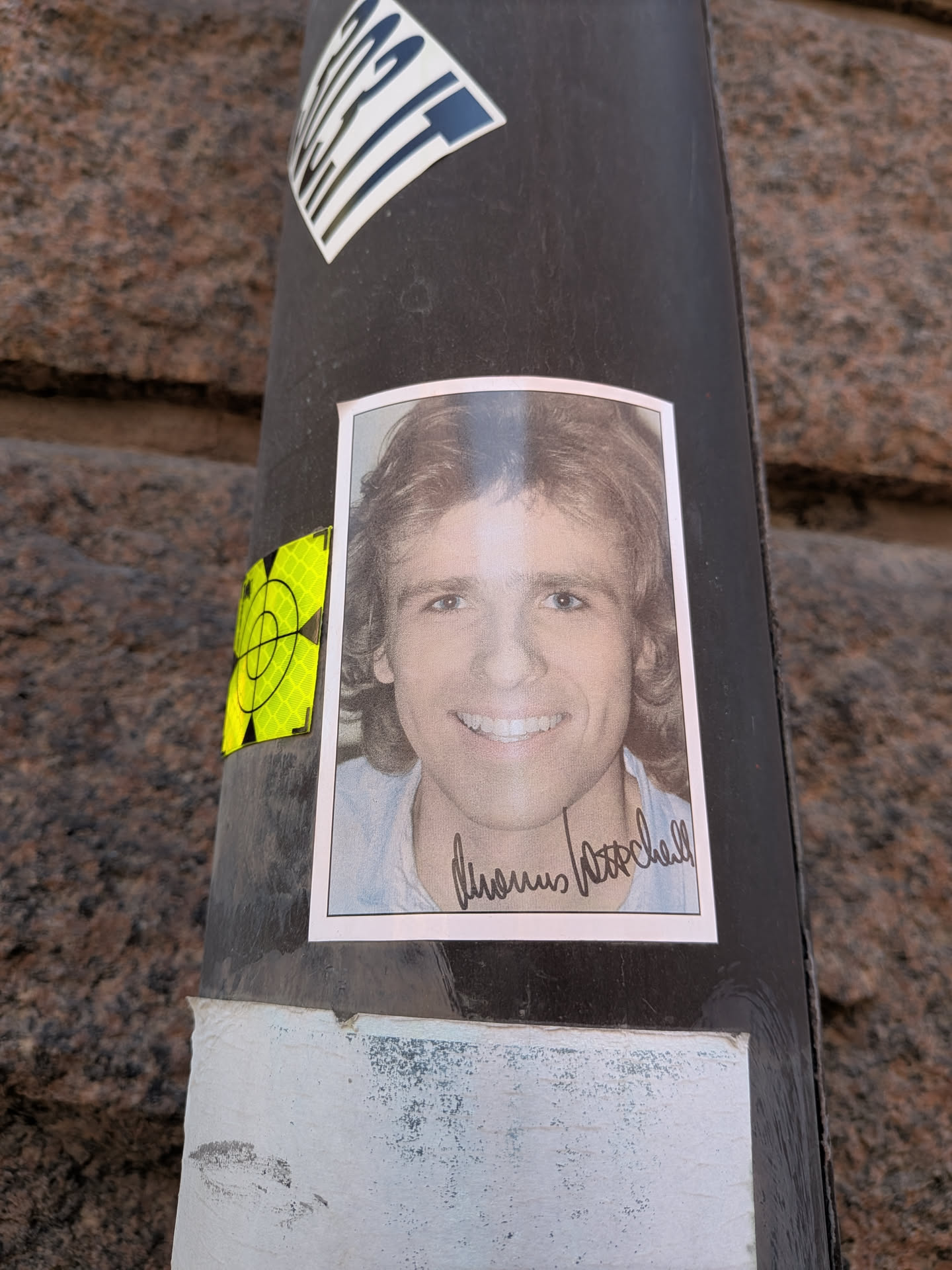 The sticker features a rectangular portrait of a smiling man with light brown, wavy, shoulder-length hair, styled in a retro fashion. He has blue eyes and a wide, friendly smile, showing his teeth. He appears to be wearing a light-colored, possibly blue, collared shirt. Below his chin, a dark, handwritten signature is prominently displayed. The sticker has a thin white border around the photographic image and is adhered to a dark pole.