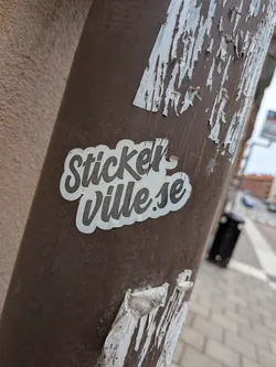 Street sticker A die-cut vinyl sticker with a white background and dark grey cursive lettering that spells out 'StickerVille.se'. The sticker is placed on a brown cylindrical metal post, surrounded by peeling remnants of older paper stickers.