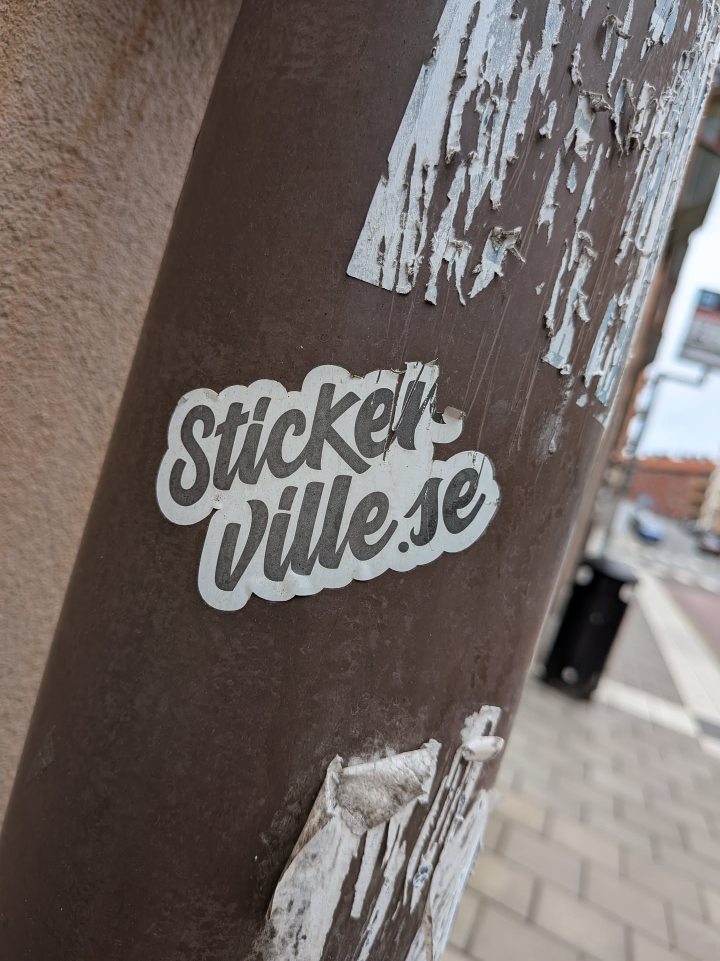 A die-cut vinyl sticker with a white background and dark grey cursive lettering that spells out 'StickerVille.se'. The sticker is placed on a brown cylindrical metal post, surrounded by peeling remnants of older paper stickers.