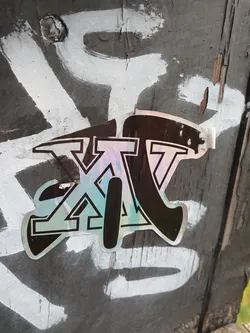 Street sticker A holographic sticker with the letters XV in a stylized design. The sticker has a dark background with a light iridescent effect.  The letters appear to have a geometric pattern incorporated into their design. The sticker is affixed to a weathered piece of wood with other graffiti markings around it.