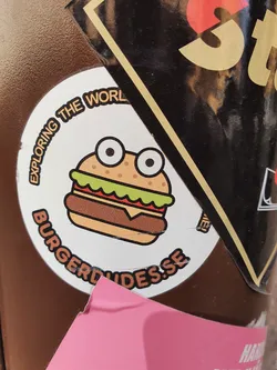 Street sticker A round sticker featuring a cartoon burger with googly eyes. The text around the burger says "Exploring the world" and "burgerguides.se".