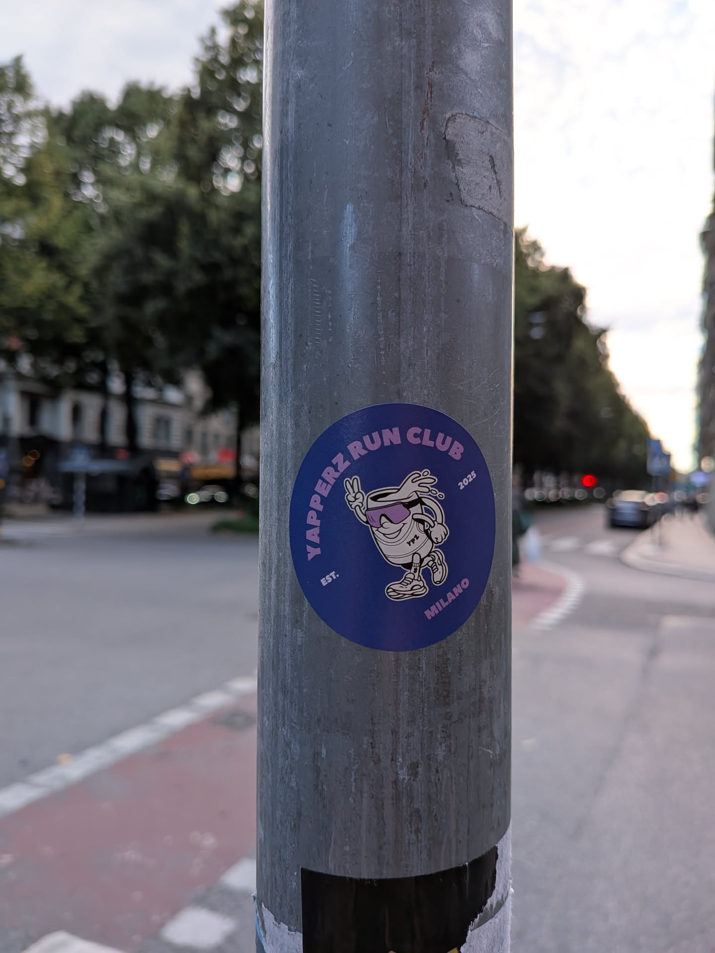 A circular sticker with a dark purple background. In the center, there is a cartoon illustration of a white, anthropomorphic cup with sunglasses and a peace sign. The cup is wearing athletic shoes and appears to be running. Purple text surrounds the illustration. 'YAPPERZ RUN CLUB' is written in an arc at the top, 'EST.' is on the left, '2025' is on the right, and 'MILANO' is at the bottom. The overall style is reminiscent of retro cartoon art.