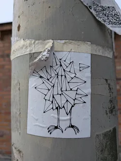 Street sticker The sticker features a minimalist, black-and-white line drawing of a bird composed of geometric shapes, resembling origami. The bird has two thin legs with clawed feet. Above and to the right of the bird, a small paper airplane is depicted. The sticker is affixed to a concrete pole and shows signs of wear, with some peeling edges.