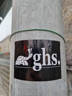 Street sticker A black square sticker with a white illustration of a turtle wearing a top hat, accompanied by the stylized letters 'ghs.' The sticker is affixed to a grey metal pole.